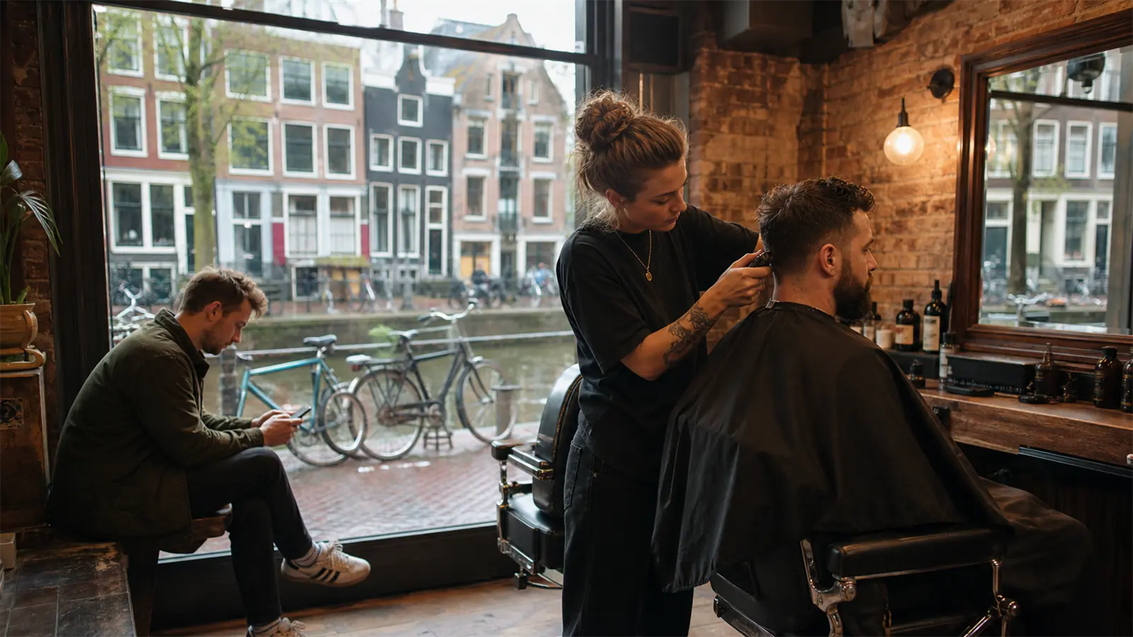 A barber cuts a client's hair in a brick-walled barbershop with a large window onto a canal and parked bicycles, another client waiting with his phone